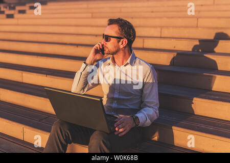 Unternehmer Gespräch am Handy und Laptop beim Sitzen auf der Treppe im Freien. Stockfoto