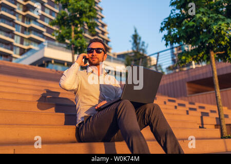 Moderne Freiberufler sprechen auf Smartphone und mit Laptop beim Sitzen auf der Treppe vor der Gebäude bei Sonnenuntergang. Stockfoto