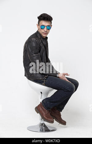 Ein Mann in einem schwarzen Lederjacke und in dunklen Spiegel Brille sitzt auf einem Stuhl, der auf einem weißen Hintergrund, ein Foto im Studio Stockfoto