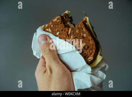Sandwich mit Käse und Schinken aus schwarzem Brot mit Getreide in die Hand eines Mannes auf einem grauen Hintergrund Stockfoto