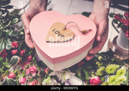 Der Florist Arbeitsplatz: Blumen, Zubehör und Werkzeugen. Weibliche Hände halten ein Geschenk in der Form eines Herzens. Stockfoto