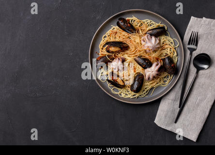 Spaghetti mit Muscheln und Tintenfisch in graue Platte mit Geschirr und Servietten auf schwarzem Hintergrund. Ansicht von oben flach, freier Speicherplatz Stockfoto