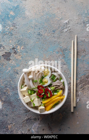 Heiße Suppe mit Nudeln, Chili, Tofu, grüne Zwiebeln und Pilze in Schale mit Stöcken auf konkrete blauen Hintergrund. Vertikal oben vie Stockfoto