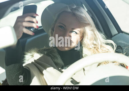 Narzisstische junge Frau Treiber ein selfie auf Ihrem Handy wie sie Laufwerke ihr Auto in der Stadt, in einer high key sonnigen Portrait Stockfoto