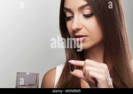 Trinken Arzneimittel. Hübsche, junge Frau, die eine schwarze Pille und hält ein Glas Wasser. Auf grauem Hintergrund. Das Konzept der Gesundheitsversorgung. Stockfoto