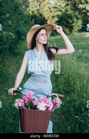 Portrait von niedlichen eine junge Frau in Hut mit Vintage Fahrrad- und Blumen auf Natur Hintergrund im Sonnenlicht im Freien. Fahrrad mit Korb voller rot und Stockfoto