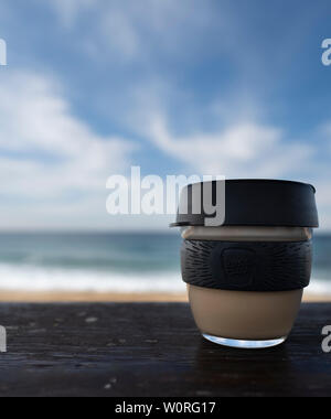 NEWCASTLE, AUSTRALIEN - 16. Juni 2019 in der Nähe von einem Glas Keepcup mit schwarzen Trimmen, gefüllt mit Kaffee, auf einer Bank mit den Pazifischen Ozean in t Stockfoto