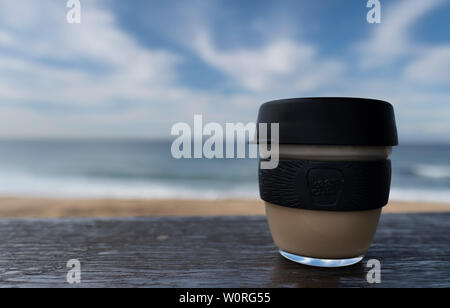 NEWCASTLE, AUSTRALIEN - 16. Juni 2019 in der Nähe von einem Glas Keepcup mit schwarzen Trimmen, gefüllt mit Kaffee, auf einer Bank mit den Pazifischen Ozean in t Stockfoto