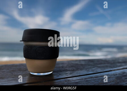 NEWCASTLE, AUSTRALIEN - 16. Juni 2019 in der Nähe von einem Glas Keepcup mit schwarzen Trimmen, gefüllt mit Kaffee, auf einer Bank mit den Pazifischen Ozean in t Stockfoto