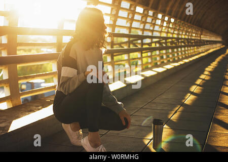 Schlanke junge brünette Frau Street Dancer gymnast sitzen und mit thermo Cup auf Street Bridge bei Sonnenaufgang Stockfoto