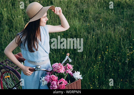 Portrait von niedlichen eine glückliche junge Frau mit Hut mit Vintage Fahrrad- und Blumen auf Natur Hintergrund im Sonnenlicht im Freien. Fahrrad mit Korb voller re Stockfoto