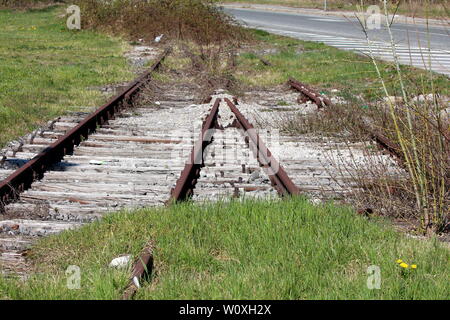 Überwucherte alte unbenutzte verrostete Gleise ruhen auf hölzernen Bahnschwellen Neben gepflasterte Straße an warmen Frühlings Stockfoto