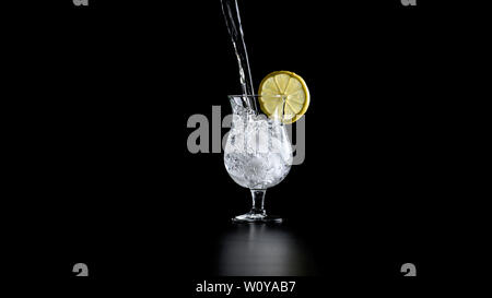 Wasser gießen in einem Cocktail Glas mit Zitronenschale auf der Kante. Über schwarzen Hintergrund. Stockfoto