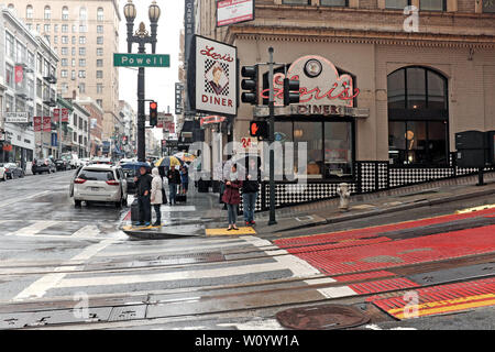 Ein Paar hält Regenschirme vor Loris Diner, während sie bei einem Regensturm in San Francisco, Kalifornien, USA die Powell Street überqueren. Stockfoto