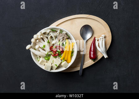 Warme japanische Nudelsuppe mit Rindfleisch, gelbe Paprika, Champignons, grüne Zwiebeln, Tofu und Chili auf schwarzem Hintergrund mit Löffel und Holz- fach Konzept der Stockfoto