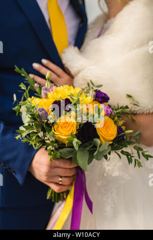 Brautstrauß für die Braut am Tag der Hochzeit. Ein schönes Mädchen hält einen Strauß gelber Rosen und lila Blüten. Der Bräutigam in einem blauen Anzug umarmt die Taille des br Stockfoto