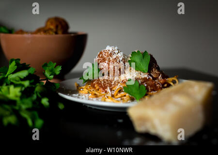 Nahaufnahme eines Tellers mit rustikalen Spaghetti und Fleischbällchen mit einer Schüssel Fleischbällchen, Parmesankäse und frischer Petersilie. Stockfoto