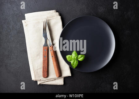 Tabelle einstellen. Schwarz Teller, Besteck, Serviette und Basilikum Blätter über schwarzen Hintergrund. Stockfoto