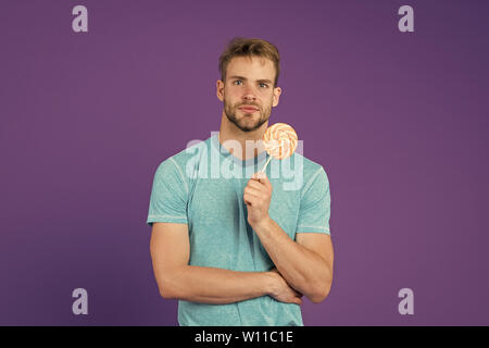 Lollipop Spaß. Mann essen großen bunten süßen Lutscher. Mann mit Borsten mag Lollipop. Cheat meal Konzept. Zucker schädlich für die Gesundheit. Kerl halten Lollipop candy violett unterlegt. Geschmack der Kindheit. Stockfoto