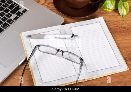 Nahaufnahme eines geöffneten Notebook, Brillen und Stift auf ein Schreibtisch mit Laptop ond Tasse Kaffee im Hintergrund