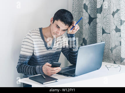Portrait des jungen Mannes in Freizeitkleidung auf dem Job Arbeiten am Laptop, Smartphone und mit nachdenklichen Gesicht Stockfoto
