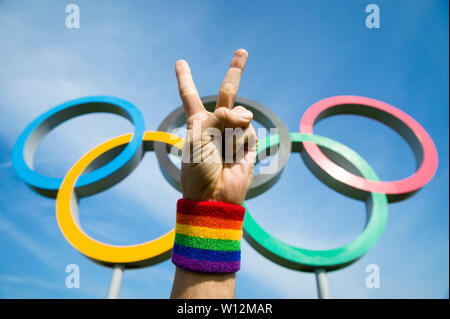 LONDON - Mai 4, 2019: eine Hand tragen stolz LGBTQI regenbogenfarbenen Armband macht einen Frieden Zeichen vor der olympischen Ringe unter blauem Himmel stehen. Stockfoto