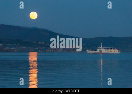 Vollmond über einige Hügel am Trasimeno See, perfekt auf dem Wasser mit einer Fähre, die in der Nähe Stockfoto