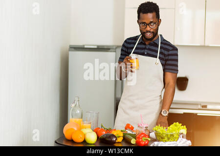 Schuß von gutaussehenden Jungen dunkelhäutigen Mann in Brillen holding Glas mit frisch gepresstem Orangensaft, bei schmackhaften Drink in leichter Küche zu Hause in Stockfoto