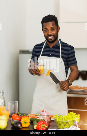 Attraktive fröhliche glückliche junge Bartgeier afrikanischen Mann hält ein Glas frisch gepressten O-Saft oder Smoothie und Messer, die Vorbereitung von pflanzlichen sa Stockfoto