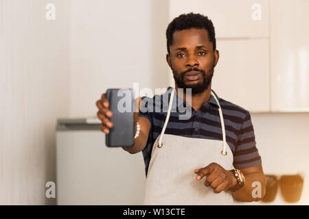 Nahaufnahme von attraktiven Bärtigen afrikanischen amerikanischen Jungen Mann Schürze mit leeren schwarzen Bildschirm stehen in der Küche smartphone. Selektive Schwerpunkte Stockfoto