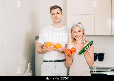 Schöne und glückliche Junge kaukasier Paar zusammen vorbereiten Abendessen in der Küche. Blonde Frau in den Händen halten, Gurken, Tomaten, ihr Ehemann holdin Stockfoto