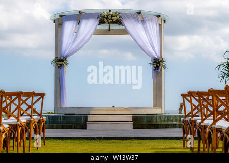 Hochzeit Bogen für eine Zeremonie mit weißem Stoff und Rosen dekoriert. Dunkle hölzerne Stühle. Blauer Himmel und Wolken im Hintergrund. Tropische Hochzeit. Stockfoto