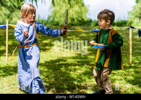 Zwei Kinder, Junge, 4-5 Jahre alt, und ein Mädchen, 5-6 Jahre alt, in Mittelalterliche Kleidung und spielerisch den Kampf mit Schwert und Axt auf Gras. Stockfoto