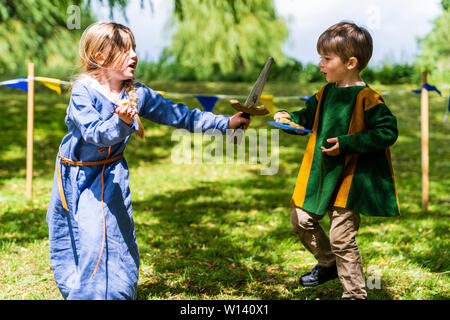 Zwei Kinder, Junge, 4-5 Jahre alt, und ein Mädchen, 5-6 Jahre alt, in Mittelalterliche Kleidung und spielerisch den Kampf mit Schwert und Axt auf Gras. Stockfoto