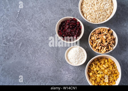 Zutaten für die Zubereitung von gesunder Bio Energie Kugeln - Nussbaum, Haferflocken, Rosinen, getrocknete Cranberries, Kokos Späne auf grauen Hintergrund, Flach, Stockfoto