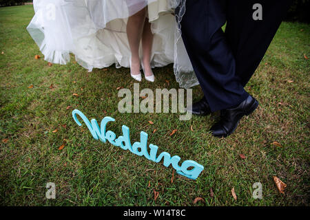 Die Beine des Bräutigams in schwarze Schuhe und Bräute in weißen Heels mit einer schneeweißen Saum von einem Hochzeitskleid auf grünem Gras, neben dem Buchstaben Hochzeit. Stockfoto