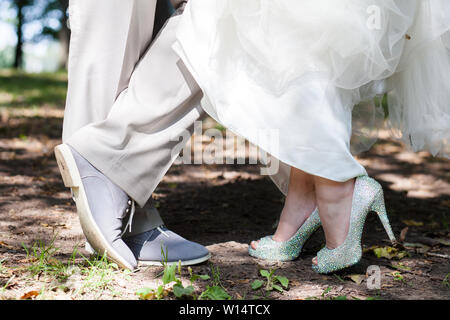 Die Beine des Bräutigams in grau Schuhe und Hose und Bräute in weiß mit Strass Heels mit einer schneeweißen Saum ein Brautkleid. Außenaufnahme Stockfoto