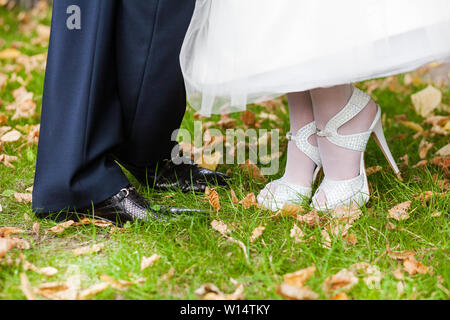 Die Beine der Bräutigam in schwarze Schuhe und Hose. Die Braut in weiß mit Strass Heels mit einer schneeweißen Saum ein Hochzeit Kleid auf dem Gras Stockfoto