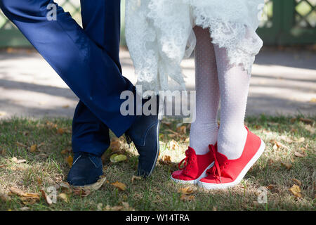 Braut in rote Schuhe mit Schnürung und Strümpfe mit einer schneeweißen Saum ein Hochzeit Kleid auf dem Gras. Die Beine des Bräutigams in blau Schuhe und Hose. Stockfoto