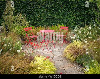 Abgeschiedenen Garten mit kleiner Terrasse und zwei roten Stühlen mit Metall Tisch von Sommer Blumen und Gräsern umgeben. Stockfoto
