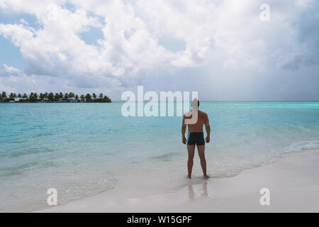 Ein starker junger Kerl steht auf einem weißen Strand und sieht auf. Im Hintergrund, riesige chic Wolken und türkisblauem Wasser. Stockfoto