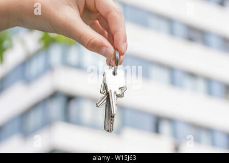 Eine Hand hält den Schlüssel zu einem gekauften Wohnung vor dem Hintergrund eines mehrstöckigen Gebäudes und grüne Blätter eines Baumes in der Gegend. Stockfoto