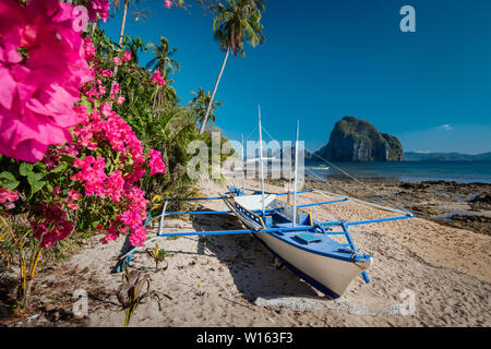 Native banca Boot und lebendigen Blumen im Las Cabanas am Strand mit erstaunlichen Pinagbuyutan Insel im Hintergrund. Exotische Natur Landschaft in El Nido, Palawan Stockfoto