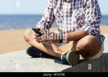 Den mittleren Abschnitt Porträt des zeitgenössischen jungen Mann mit Smartphone sitzen auf konkrete Block vom Meer aus, kopieren Sie Platz Stockfoto