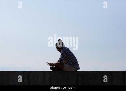 Seitenansicht Porträt des zeitgenössischen jungen Mann Chillen am Strand hören von Musik während der Sitzung auf konkrete Block gegen den blauen Himmel beschrieben, Kopieren spac Stockfoto