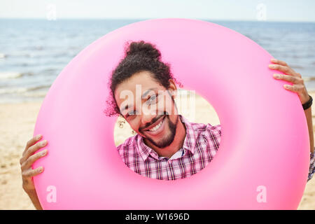 Portrait von jungen lateinamerikanischen Mann lächelt glücklich an der Kamera, während am Strand mit rosa schwimmen Rohr, Spaß Sommer Konzept posing Stockfoto