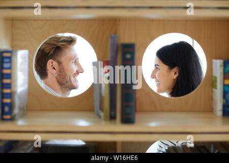 Lächelnd schöne romantische Paar an jedem anderen Suchen in der Bibliothek, Gesichter von Menschen durch runden Bücherregal Löcher Stockfoto