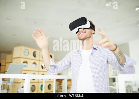 Jolly aufgeregt tätowiert Kerl mit Stoppeln legere Shirt stehen in der modernen Bibliothek und mit Virtual reality Simulator beim Spielen video game Stockfoto