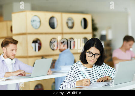 Ernste nachdenkliche junge Frau in Brillen am Tisch sitzen und notieren Sie sich die Informationen im Notebook Stockfoto