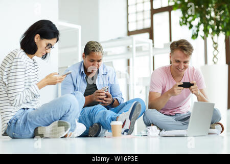 Gruppe von Technologie-süchtigen Studenten in Freizeitkleidung sitzt entspannt Posen auf dem Boden und mit modernen Geräten, während für die Suche nach Informationen in Stockfoto